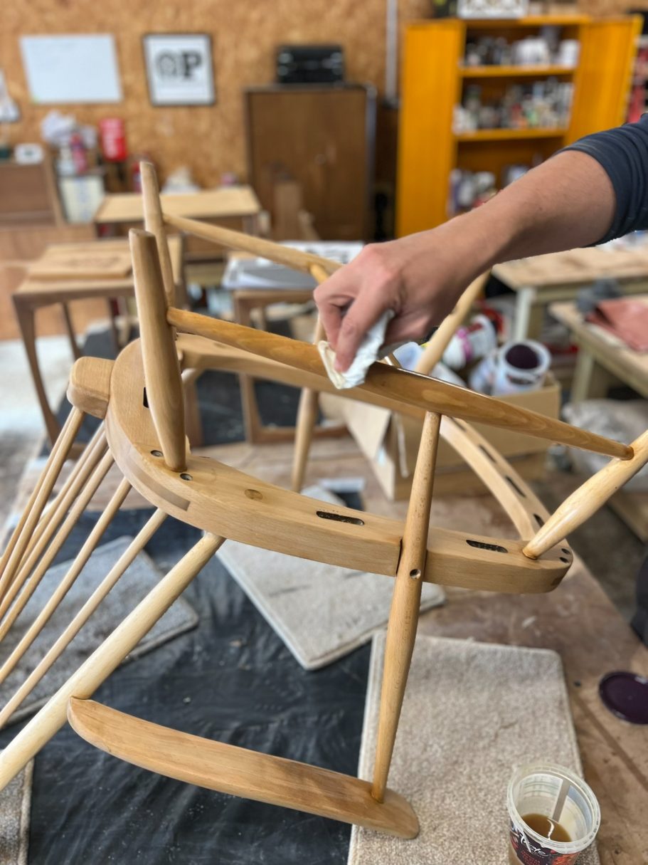 Applying the oil A person refinishes a wooden chair with a cloth in a workshop setting.